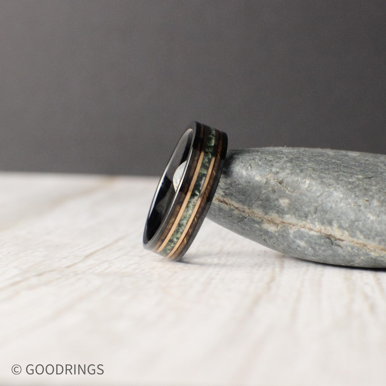 May include: A black tungsten carbide ring with a wood inlay. The inlay features green and brown wood with a black line separating the two colours.