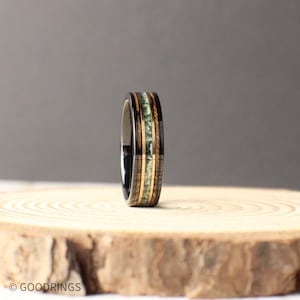 May include: A black tungsten carbide ring with a band of green and brown wood inlay. The ring is on a wooden surface.