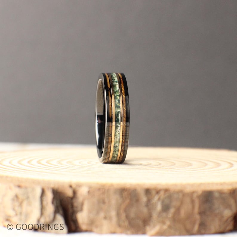 May include: A black tungsten carbide ring with a band of green and brown wood inlay. The ring is on a wooden surface.