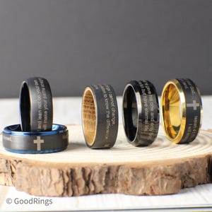 May include: A collection of engraved rings with religious inscriptions and a cross design. The rings are displayed on a wooden slice, showcasing various colors including black, blue, and gold. The text includes phrases like "Isaiah 41:10" and "eternal life."