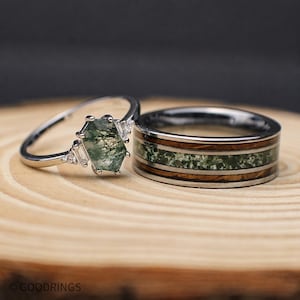 May include: A silver-toned ring featuring a hexagonal moss agate stone flanked by small baguette and round clear stones, alongside a wider silver-toned band with a central inlay of moss agate and wood grain, both marked with the text GOODRINGS.