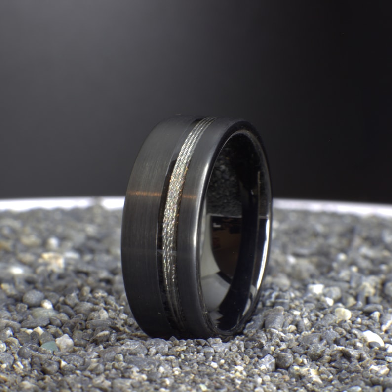 May include: A black tungsten carbide ring with a silver inlay. The ring has a brushed finish and a polished edge.