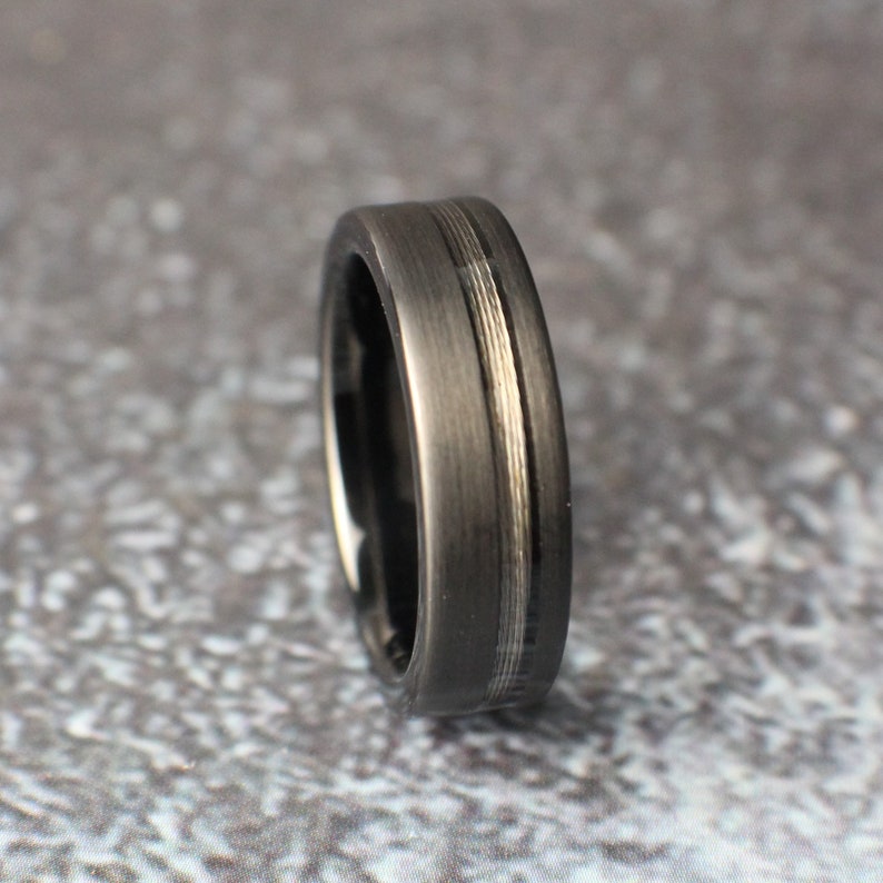 May include: A black tungsten carbide ring with a brushed finish and a silver inlay. The ring is a classic style and would make a great wedding band.