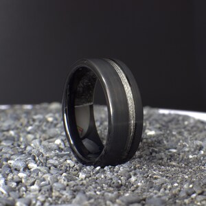 May include: A black tungsten carbide ring with a silver inlay. The ring has a brushed finish and a beveled edge.