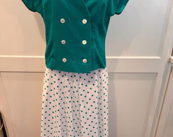 Könnte beinhalten: Ein zweiteiliges Vintage-Outfit. Das tealgrüne Oberteil hat kurze Ärmel, einen V-Ausschnitt und vier weiße Knöpfe. Der Rock ist weiß mit tealgrünen Polka Dots. Das Outfit wird auf einer Schaufensterpuppe präsentiert. Stil der 80er Jahre.