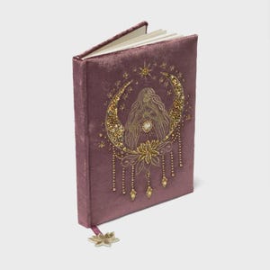Könnte beinhalten: Ein Notizbuch aus altrosa Samt mit goldener Paillettenstickerei auf dem Einband. Das Design zeigt eine zentrale Figur, umrahmt von einer Mondsichel, Sternen und hängenden Perlenakzenten. Ein passender goldener Lotos-Anhänger hängt am Buchrücken.