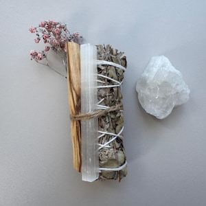 Könnte beinhalten: Ein Bündel getrockneter Salbei, Palo Santo und ein Selenit-Kristallstab, gebunden mit Schnur und weißem Faden, neben einem rohen Quarz-Kristall und rosa Trockenblumen. Auf einer grauen Oberfläche angeordnet.