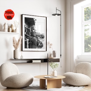 May include: A black and white framed photograph of a marketplace scene hangs on a white wall. The room features a modern design with a round coffee table, two beige armchairs, and a black floor lamp. Decorative vases and plants add to the decor.