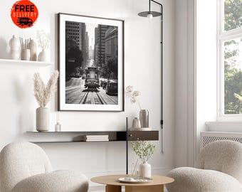 May include: Black and white photograph of a streetcar in San Francisco, California. The streetcar is in the middle of the street, with buildings on either side. The photograph is framed and hanging on a white wall.