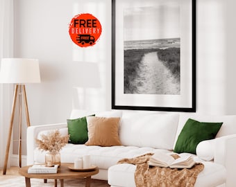 May include: A black and white photograph of a path leading to the ocean. The path is surrounded by tall grass and the ocean is in the background. The photo is framed in a black frame and hanging on a white wall above a white couch with green and brown throw pillows.