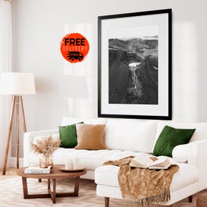 May include: A black and white framed landscape photograph hangs above a white sectional sofa with green and tan throw pillows. A wooden coffee table holds a vase of dried flowers and a book. A floor lamp stands to the left.