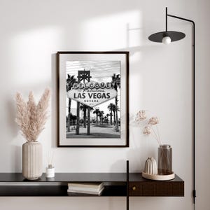 May include: Black and white framed print of the iconic "Welcome to Fabulous Las Vegas" sign, with palm trees in the background. The sign is centered in the image, with the text "Las Vegas Nevada". The print is displayed on a white wall.