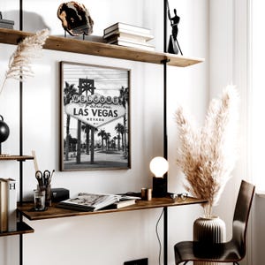 May include: A black and white framed print of the iconic "Welcome to Fabulous Las Vegas Nevada" sign. The image is displayed on a wooden desk with a lamp, books, and decorative items. The scene is a modern office or home decor setting.