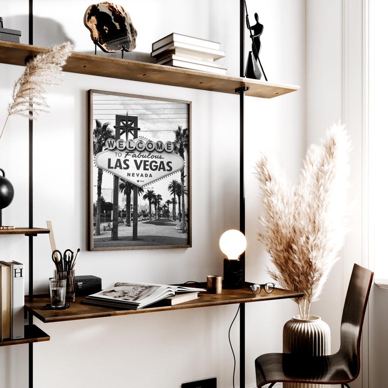 May include: A black and white framed print of the iconic "Welcome to Fabulous Las Vegas Nevada" sign. The image is displayed on a wooden desk with a lamp, books, and decorative items. The scene is a modern office or home decor setting.