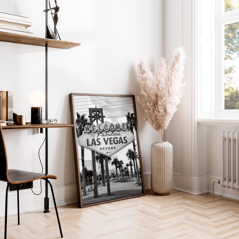 May include: A framed black and white photograph of the iconic "Welcome to Fabulous Las Vegas Nevada" sign. The image captures the sign's design with palm trees and a desert landscape. The photograph is displayed in a room with a wooden floor.
