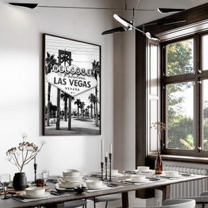 May include: Black and white framed print of the iconic "Welcome to Fabulous Las Vegas" sign, Nevada. The image is displayed in a dining room setting, with a long wooden table set for a meal. The room has a modern design with a large window.