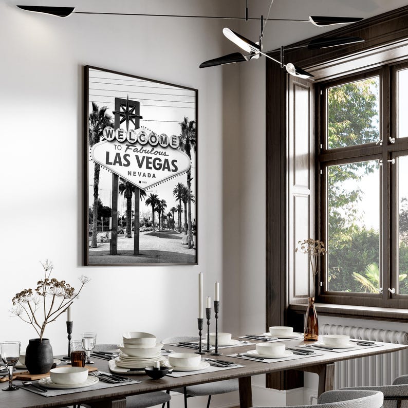 May include: Black and white framed print of the iconic "Welcome to Fabulous Las Vegas" sign, Nevada. The image is displayed in a dining room setting, with a long wooden table set for a meal. The room has a modern design with a large window.