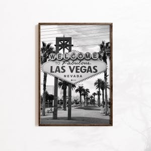 May include: Black and white framed print of the iconic "Welcome to Fabulous Las Vegas Nevada" sign. The sign is oval-shaped with a starburst design at the top and is surrounded by palm trees. The image is a classic representation of Las Vegas.