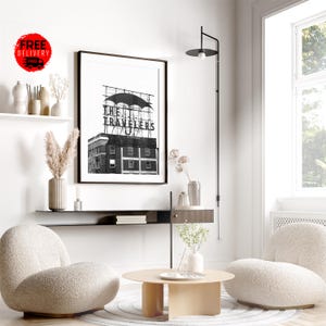 May include: A black and white framed print of a vintage sign that reads "THE TRAVELERS". The image is displayed in a modern living room setting with neutral-toned furniture, including two cream-colored armchairs and a round coffee table.