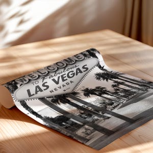 May include: Black and white print of the iconic "Welcome to Fabulous Las Vegas" sign, featuring palm trees and the city skyline. The print is rolled up on a wooden surface, showcasing the sign's classic design and the Nevada location.