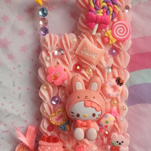 May include: A pink phone case decorated with a Hello Kitty character wearing a bunny costume, a pink and yellow lollipop, a pink cake with red cherries, and other pink and white candy decorations. The case is covered in pink frosting and has a variety of embellishments, including rhinestones, pearls, and glitter.