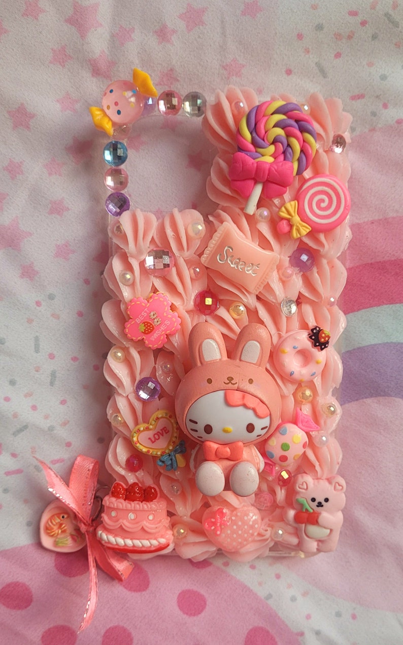 May include: A pink phone case decorated with a Hello Kitty character wearing a bunny costume, a pink and yellow lollipop, a pink cake with red cherries, and other pink and white candy decorations. The case is covered in pink frosting and has a variety of embellishments, including rhinestones, pearls, and glitter.
