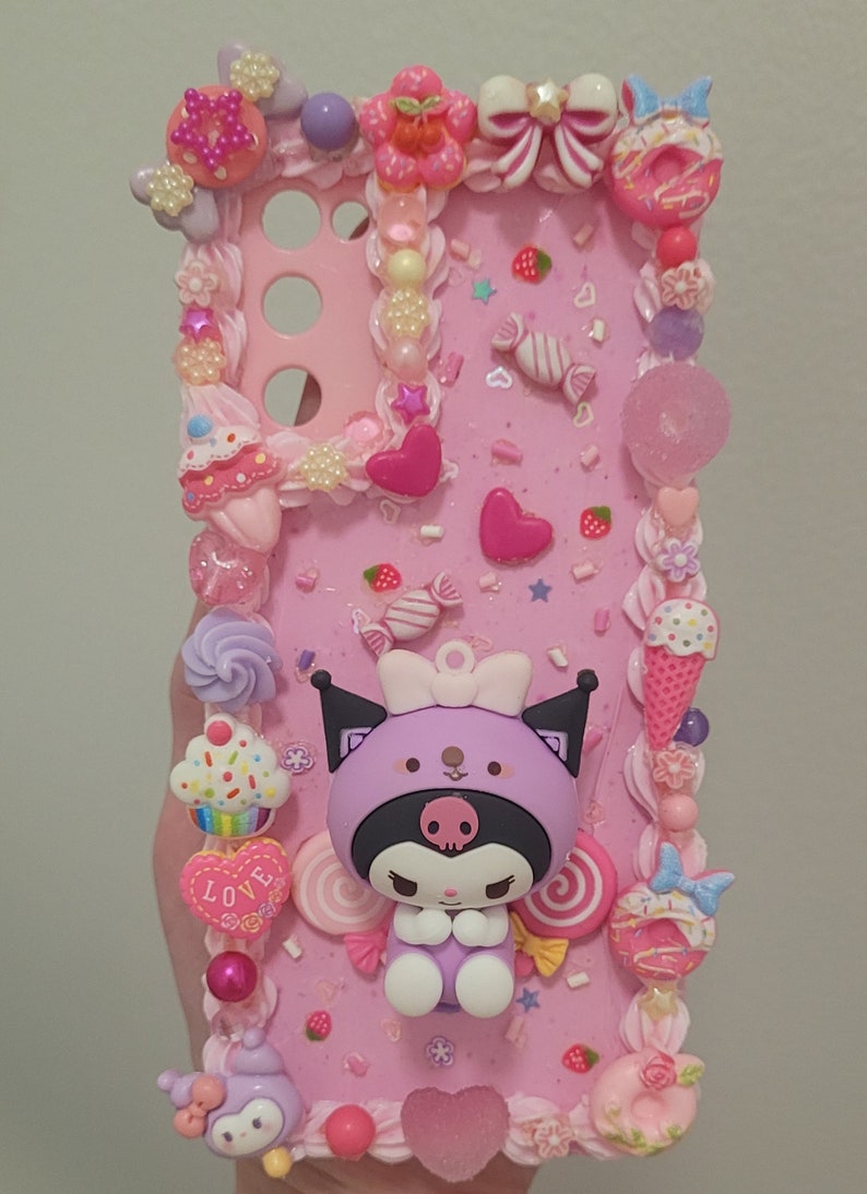 May include: A pink phone case decorated with a purple and black Kuromi character, candy, hearts, and sprinkles. The case has a raised 3D design and is made of resin.