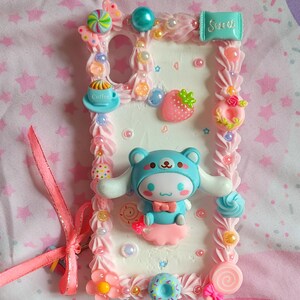 May include: A white phone case decorated with pink frosting, sprinkles, and a blue cartoon character. The case also features a small cup of coffee, a strawberry, a donut, and a lollipop.