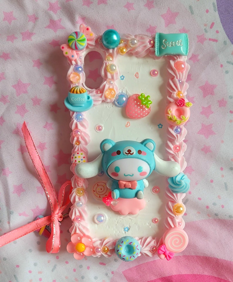 May include: A white phone case decorated with pink frosting, sprinkles, and a blue cartoon character. The case also features a small cup of coffee, a strawberry, a donut, and a lollipop.