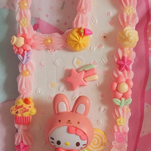 May include: A pink and white phone case decorated with a Hello Kitty character dressed as a bunny, a cupcake with the words "Sweet Cakes", a donut, a candy, and other colorful embellishments.