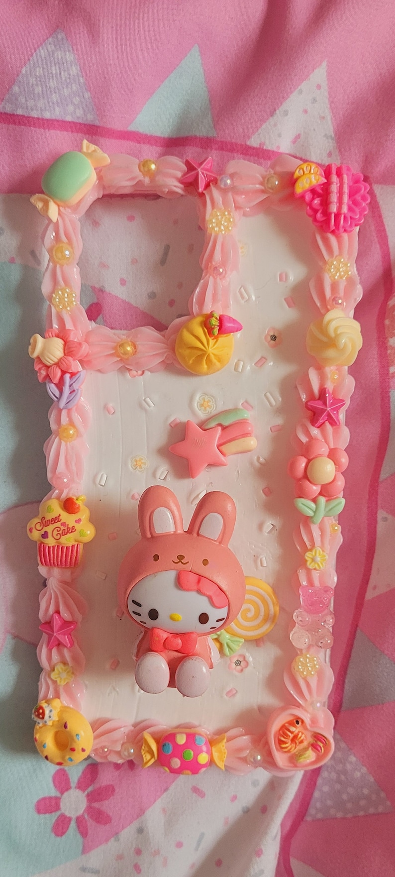May include: A pink and white phone case decorated with a Hello Kitty character dressed as a bunny, a cupcake with the words "Sweet Cakes", a donut, a candy, and other colorful embellishments.