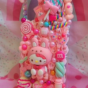 May include: A pink phone case decorated with a Hello Kitty character, candy, and other cute embellishments. The case is covered in pink, white, and blue colors.