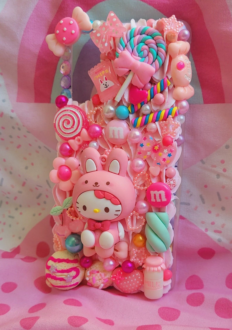 May include: A pink phone case decorated with a Hello Kitty character, candy, and other cute embellishments. The case is covered in pink, white, and blue colors.