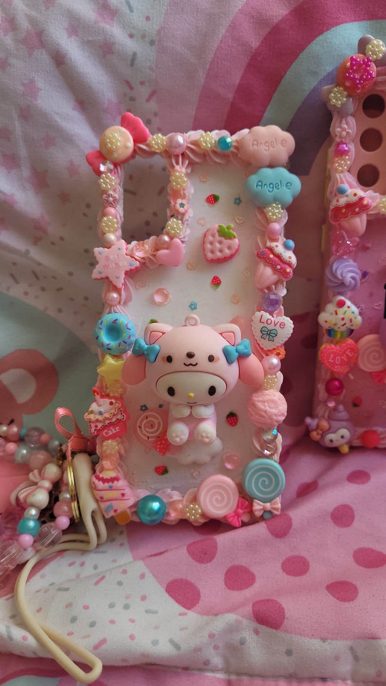 May include: A pink and white phone case decorated with a cartoon character, candy, and hearts. The case has the words "Angel-e" and "Love" on it.
