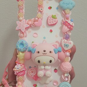 May include: A pink and white phone case decorated with a pink cartoon character, candy, and hearts. The case has the words "Love", "Cake", and "Angele" on it.