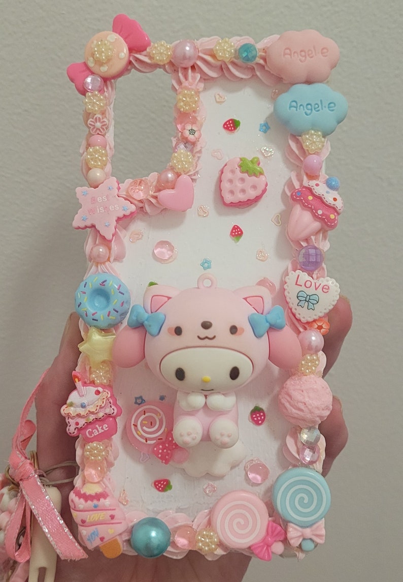 May include: A pink and white phone case decorated with a pink cartoon character, candy, and hearts. The case has the words "Love", "Cake", and "Angele" on it.