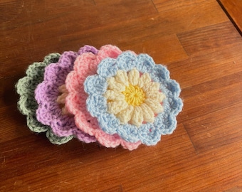 May include: A collection of five handmade crochet flower coasters in various pastel colors: green, purple, pink, light blue, and yellow. The coasters are arranged on a wooden surface.