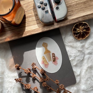 May include: A wooden tray holds a candle in a glass jar, a dried citrus slice, and a pair of silver and red game controllers. A gray notebook with a sticker featuring a raised fist and game controllers is also present.