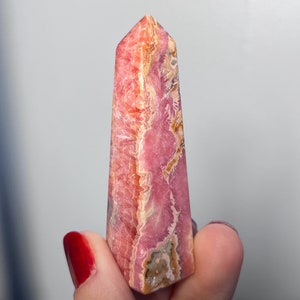 May include: A pink and white crystal point with a striated pattern. The crystal is shaped like a pyramid and has a smooth, polished surface.
