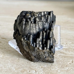 Puede incluir: Espécimen mineral oscuro y metálico con una superficie rugosa y texturizada. El mineral tiene un color gris oscuro con toques de oro y plata. La muestra se exhibe sobre un soporte transparente, mostrando su formación natural única.