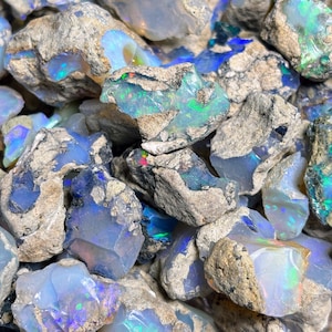 May include: A close-up of a pile of rough, gray opal stones. The stones have a variety of shapes and sizes, and some have a blue, green, or purple iridescent sheen.