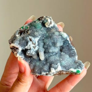 Peut inclure: Un spécimen minéral brut présentant un amas de formations cristallines gris foncé aux surfaces scintillantes et réfléchissantes. La pierre présente de petites inclusions vert vif dispersées sur la surface texturée et dentelée du rocher.