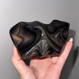 May include: A polished, irregular glass sculpture featuring a swirling, marbled pattern of deep black, charcoal gray, and subtle iridescent streaks of gold and blue. The surface is smooth and reflective, with organic, undulating curves and edges.