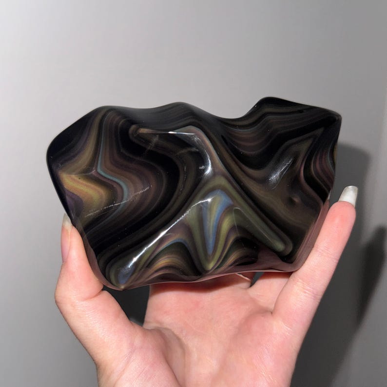 May include: A polished, irregular glass sculpture featuring a swirling, marbled pattern of deep black, charcoal gray, and subtle iridescent streaks of gold and blue. The surface is smooth and reflective, with organic, undulating curves and edges.