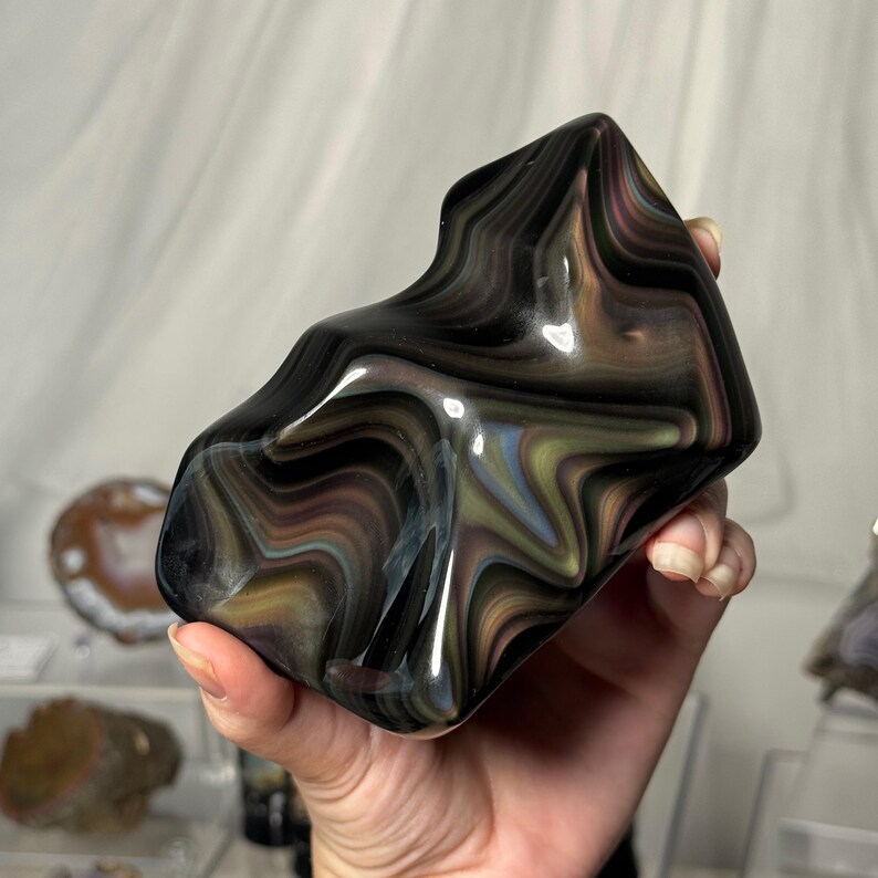 May include: A polished obsidian stone featuring a smooth, flame-like shape with swirling bands of black, iridescent gold, and subtle hints of blue and purple. The surface is highly reflective, showcasing a fluid, layered pattern throughout the piece.