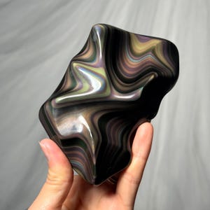 May include: A polished, irregular-shaped stone featuring a dark black base with swirling, iridescent bands of metallic green, purple, and gold colors that create a fluid, wavy pattern across the smooth, reflective surface of the mineral specimen.