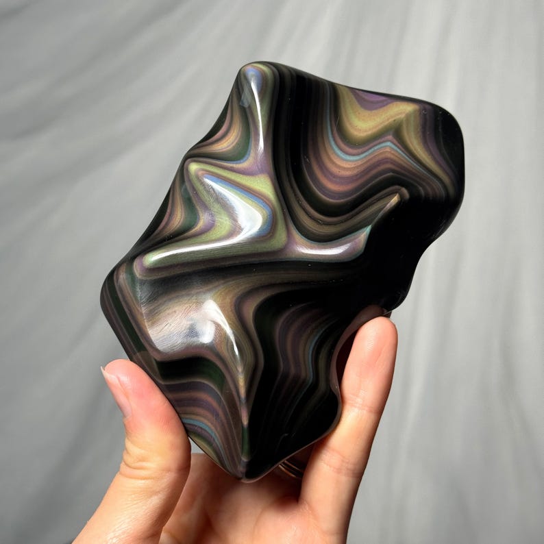 May include: A polished, irregular-shaped stone featuring a dark black base with swirling, iridescent bands of metallic green, purple, and gold colors that create a fluid, wavy pattern across the smooth, reflective surface of the mineral specimen.