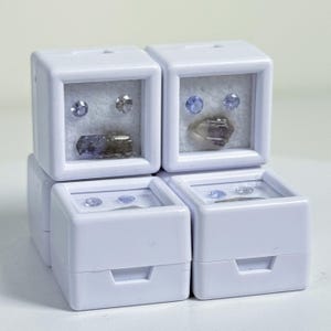 May include: Four square white plastic display cases with clear windows, each containing small loose gemstones. The stones include faceted round blue and clear crystals alongside larger, irregular raw mineral specimens in shades of blue and brown.