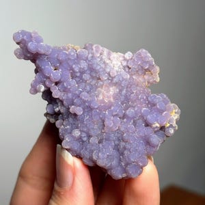 May include: A grape agate mineral specimen, displaying a striking purple colour and a distinctive bubbly, botryoidal texture. The agate is held in a hand, highlighting its natural form. The specimen's texture is made up of many small, rounded formations.