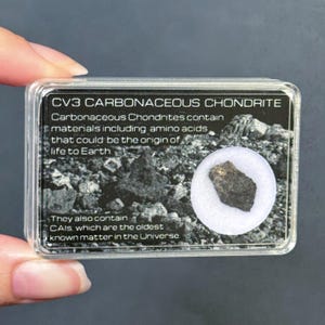 Pode incluir: Uma pequena caixa de plástico retangular contendo um pedaço de condrito carbonáceo CV3. A caixa tem um fundo preto e branco com o texto "CV3 CARBONACEOUS CHONDRITE". O fragmento de meteorito é cinza escuro e marrom.